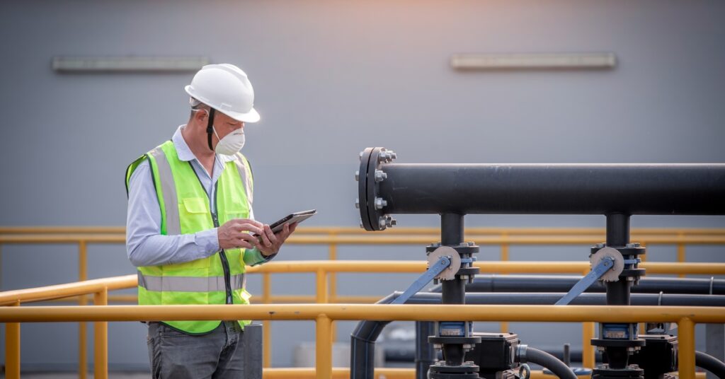 A man wearing a white hard hat, yellow safety vest, and a white face mask is holding a clipboard and observing a treatment.