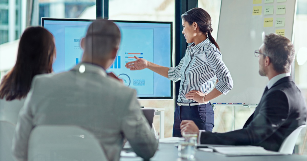 A person sits at a desk, pointing at a laptop screen that contains different graphs. Paperwork is spread around the desk.