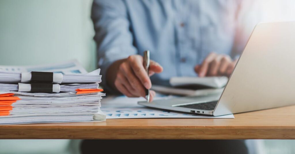 A person sits at a wooden desk typing on a calculator and holding a pen. A laptop and a pile of paperwork appears on the desk.