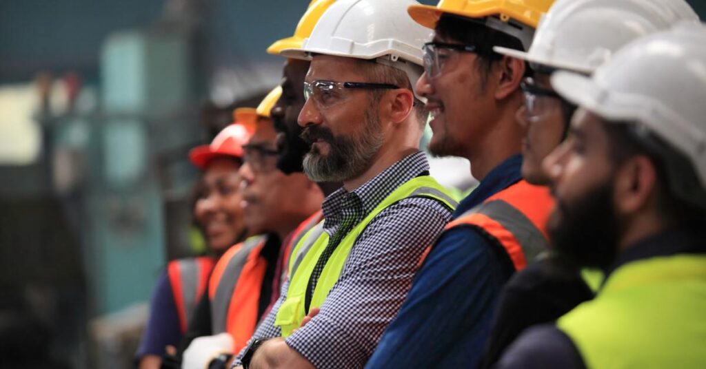 Men wearing bright orange and yellow safety vests and white and yellow hard hats stand in a line, smiling.