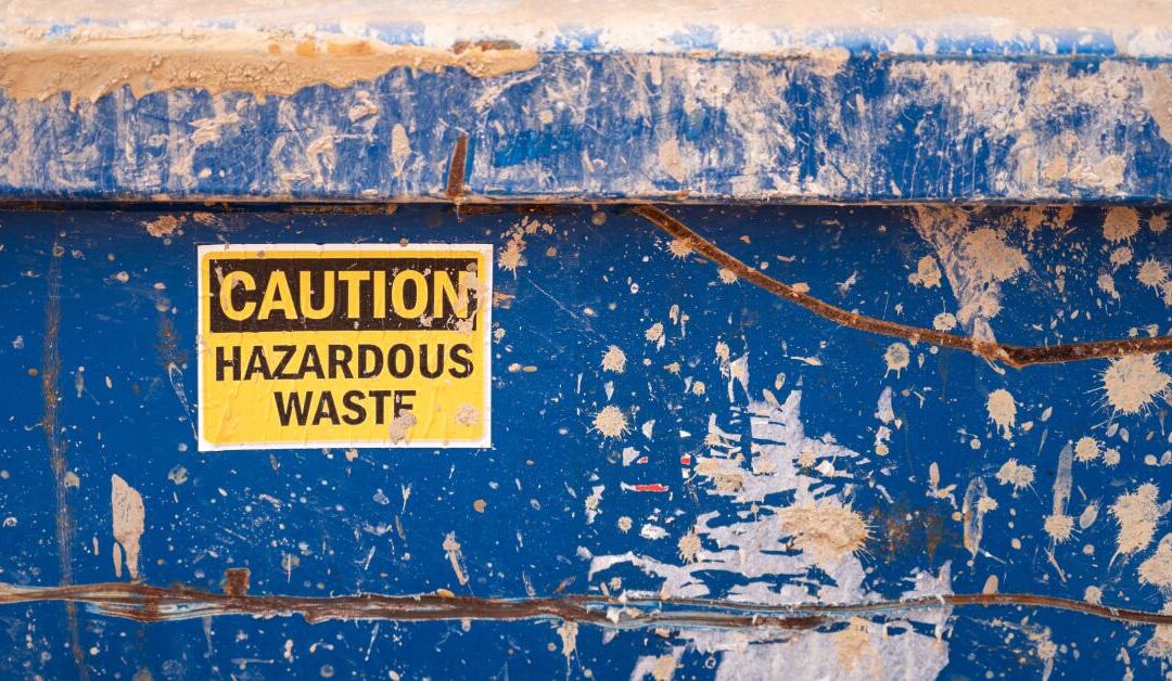 A yellow sticker on an extremely dirty surface reads "Caution Hazardous Waste" in bold, black letters.
