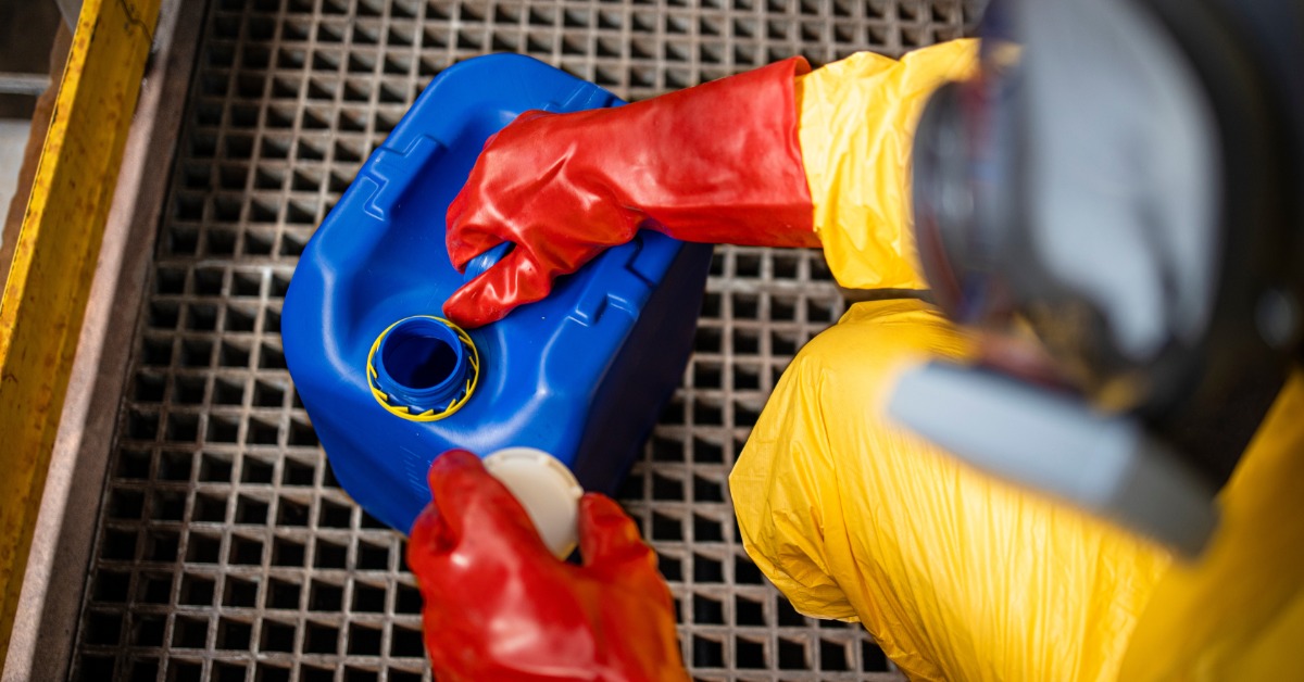 An over-the-shoulder view of a person wearing a yellow hazmat suit and red rubber gloves touches a blue container.