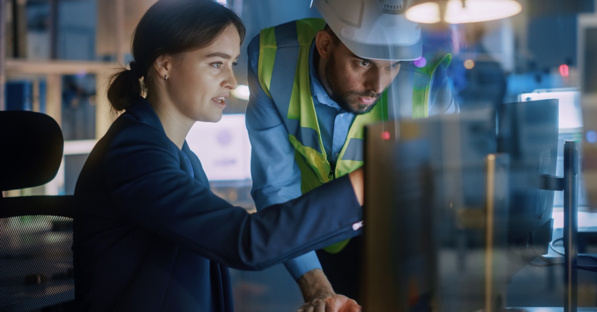 A woman sits at a desk as a man leans over the desk, looking at a desktop monitor. He is wearing a hard hat and a safety vest.
