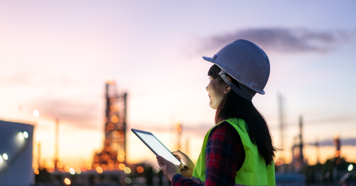 A woman wearing a white hard hat and a neon-yellow safety vest is holding a white tablet. The background is blurry.