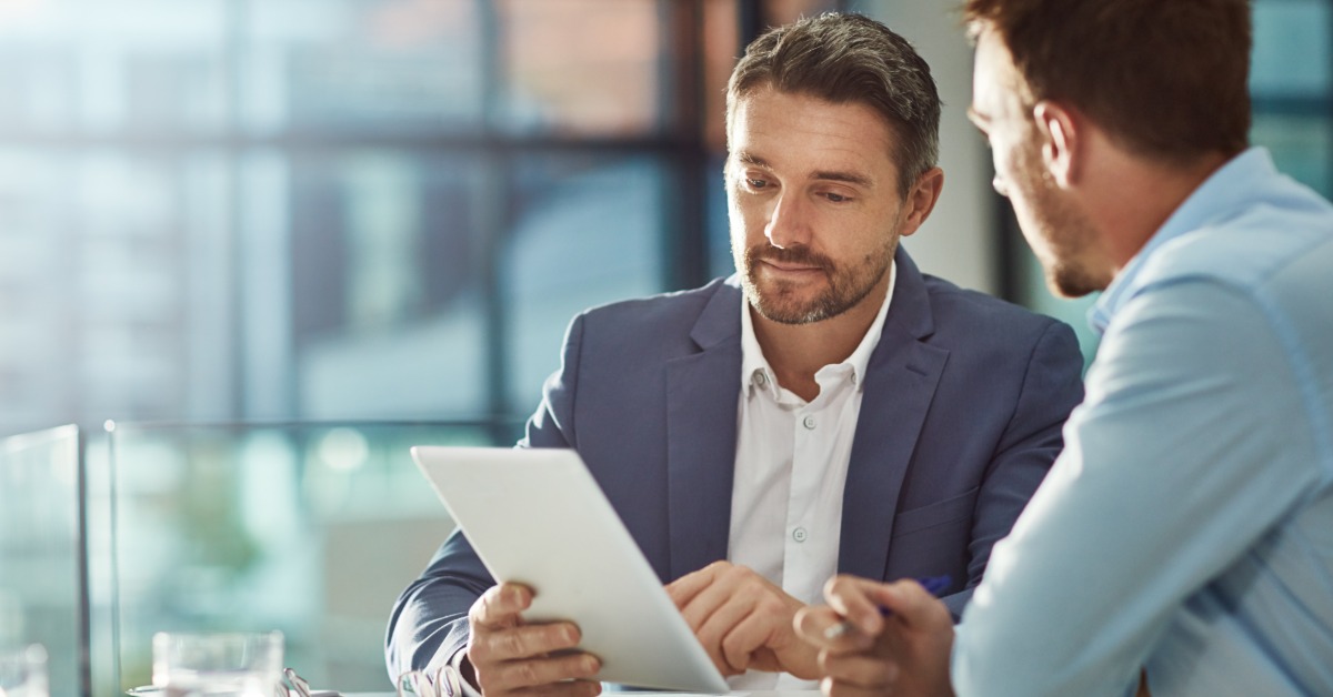 Two men wearing business attire sit at a table having a discussion. One of the men is holding a tablet.