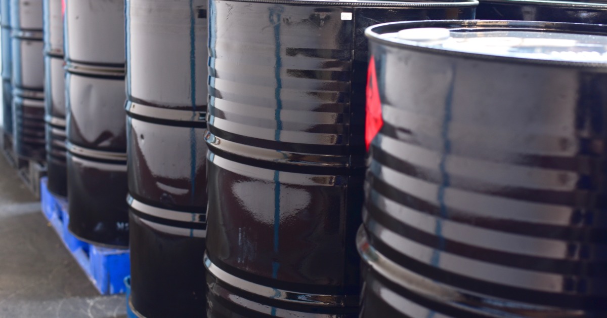 A close-up view shows a line of black, metal barrels lined up on blue pallets. The barrels are outside.
