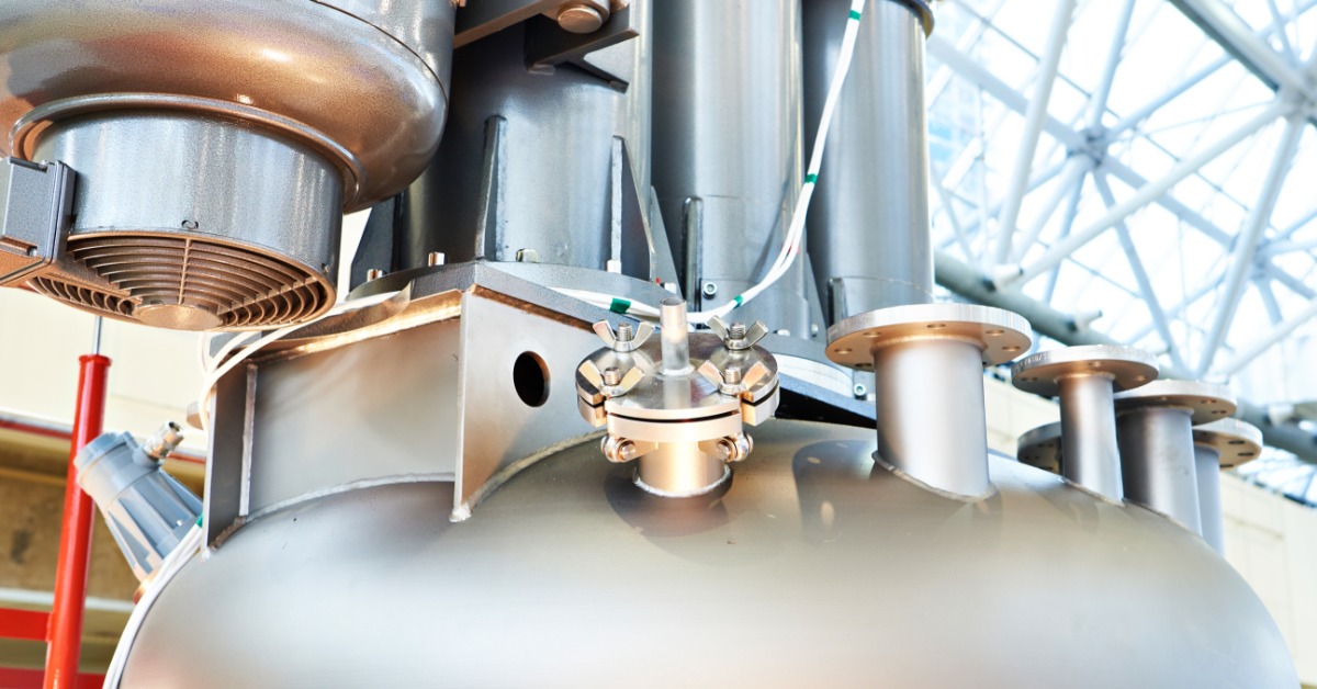 A large, metal industrial dissolver sits in an industrial warehouse. A single small, plastic tube is coming out of the dissolver.