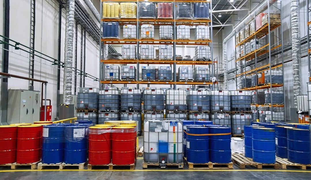 A large industrial warehouse contains several storage shelves and pallets full of drums and metal barrels.