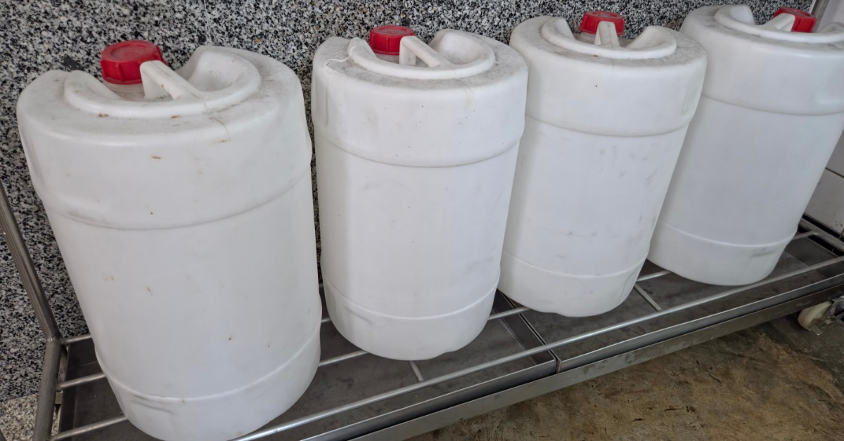 Four identical white, plastic drums contain red caps and handles and sit in a row on a metal shelving unit.