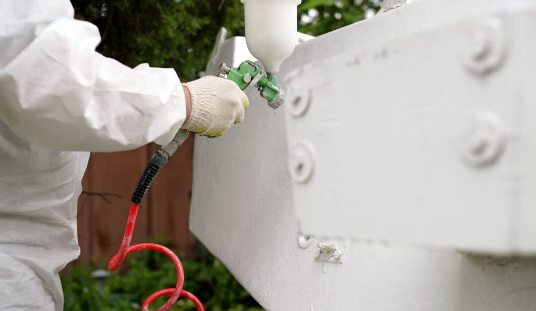 A person wearing a white protective suit is using a sprayer to spray white paint onto an item that's outside.