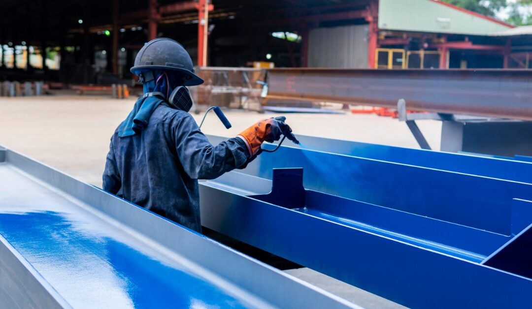 A person wearing a blue jumpsuit, a gas mask, orange work gloves, and a blue hard hat is using a sprayer to paint steel blue.