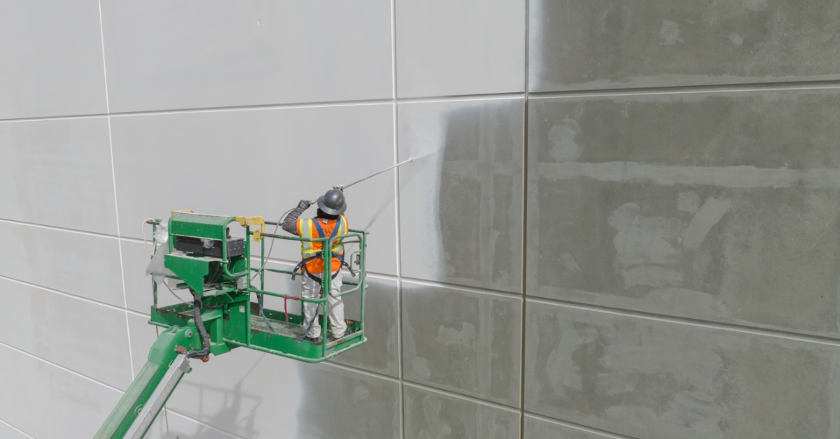 A person wearing an orange safety vest and a gray hard hat stands in a cherry picker to paint a wall with a sprayer.