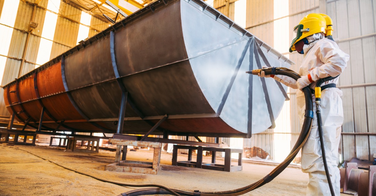 A person wearing a white protective suit and a yellow gas mask is using a sprayer to spray a large, steel industrial item.