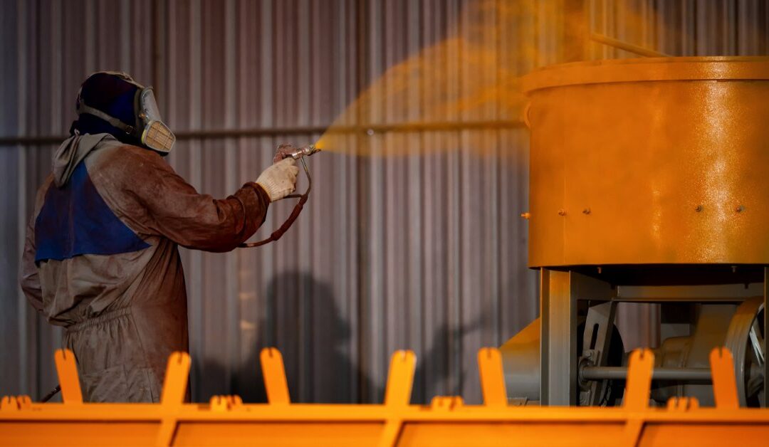 A person wearing a protective suit and a face mask is using a sprayer to paint a piece of metal orange.