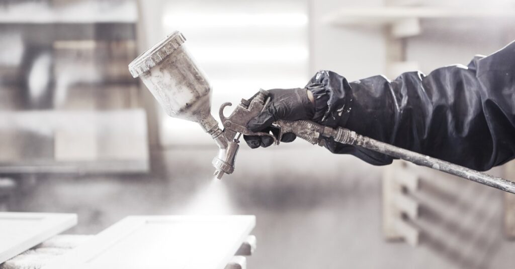 A person is wearing a black protective suit and black latex gloves as they use a sprayer to paint an object white.