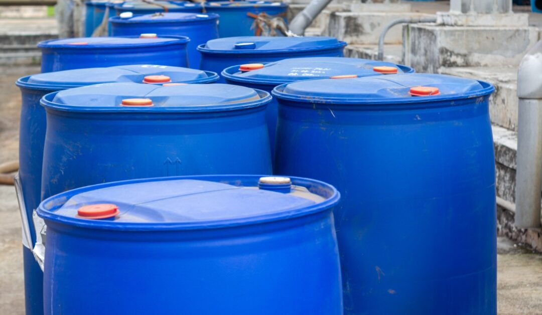 Several large, blue plastic barrels sit outside of an industrial facility in two rows. The barrels have an orange cap.