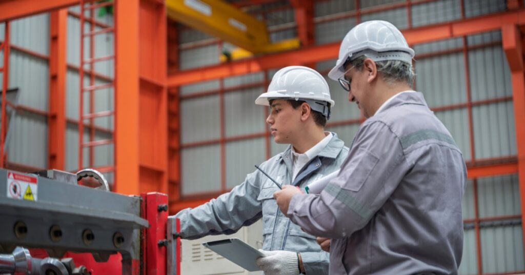 Two male team members wearing white hard hats and gray coats are standing in an industrial facility.