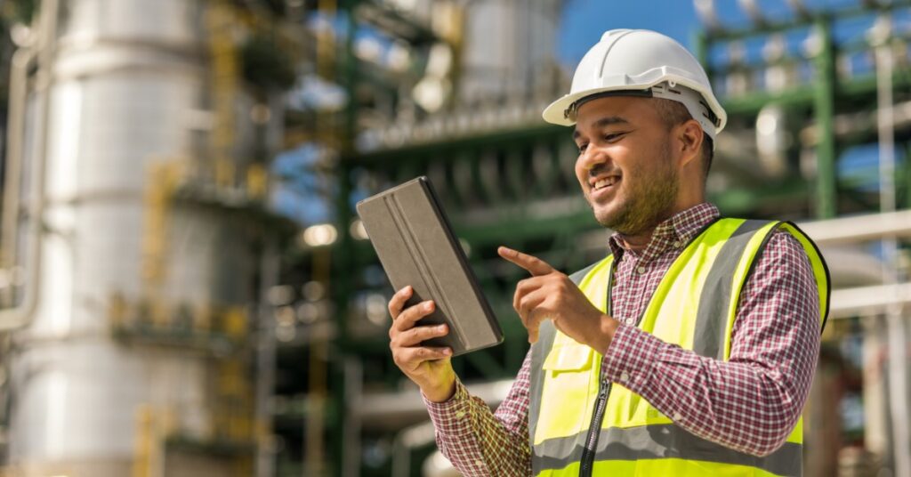 A man wearing a white hard hat and a neon-yellow safety vest stands outside of an industrial facility holding a tablet.