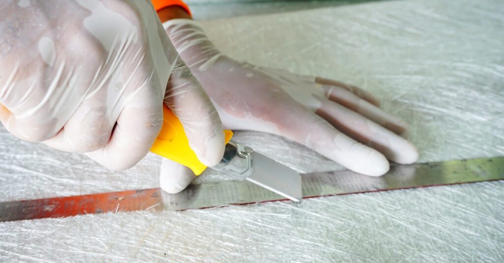 A close-up view shows a person wearing white latex gloves using an Xacto knife to cut fiberglass along the edge of a ruler.