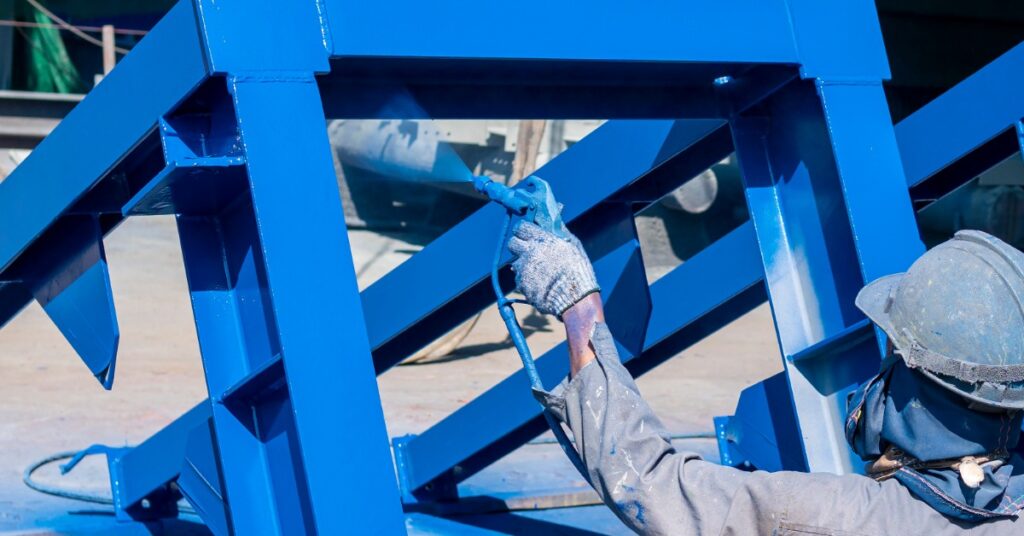 A person wearing a blue hard hat, a protective mask, gloves, and a long shirt is using a sprayer to paint metal blue.