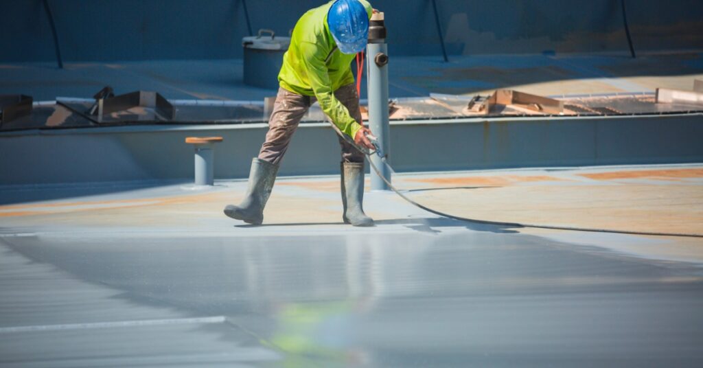 A man wearing a neon yellow shirt, rain boots, tan pants, and a blue hard hat is using a sprayer to paint concrete.