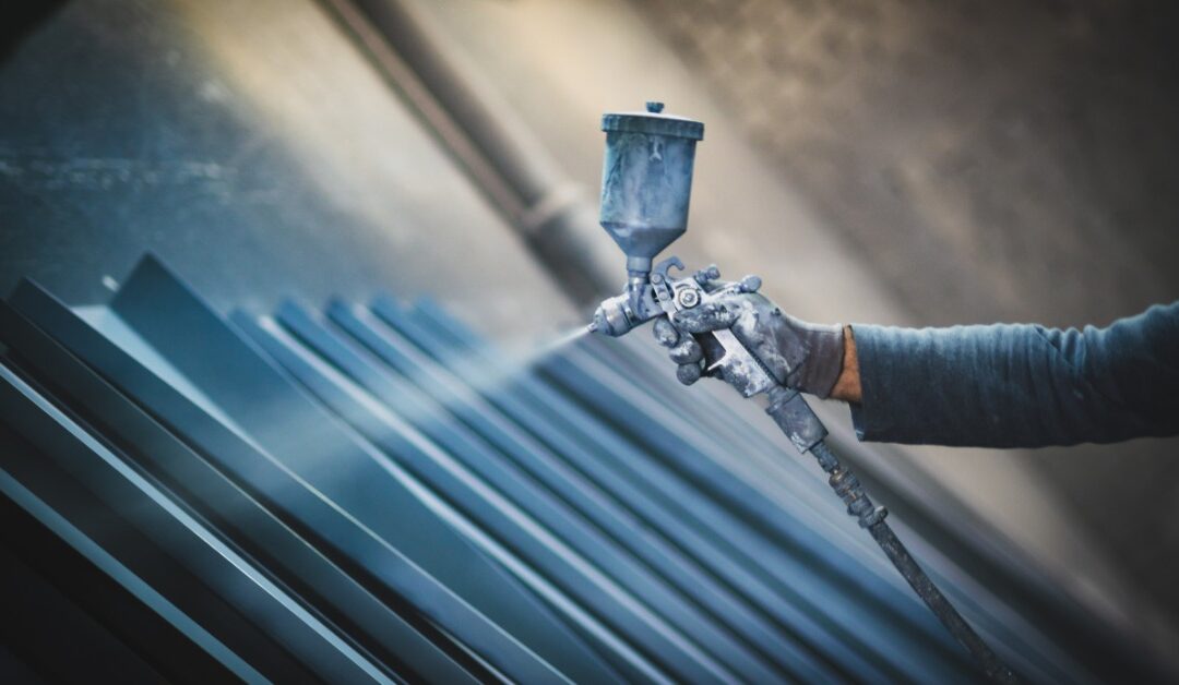 A close-up view shows a person using an industrial sprayer to spray pieces of metal. The person is wearing protective gear.