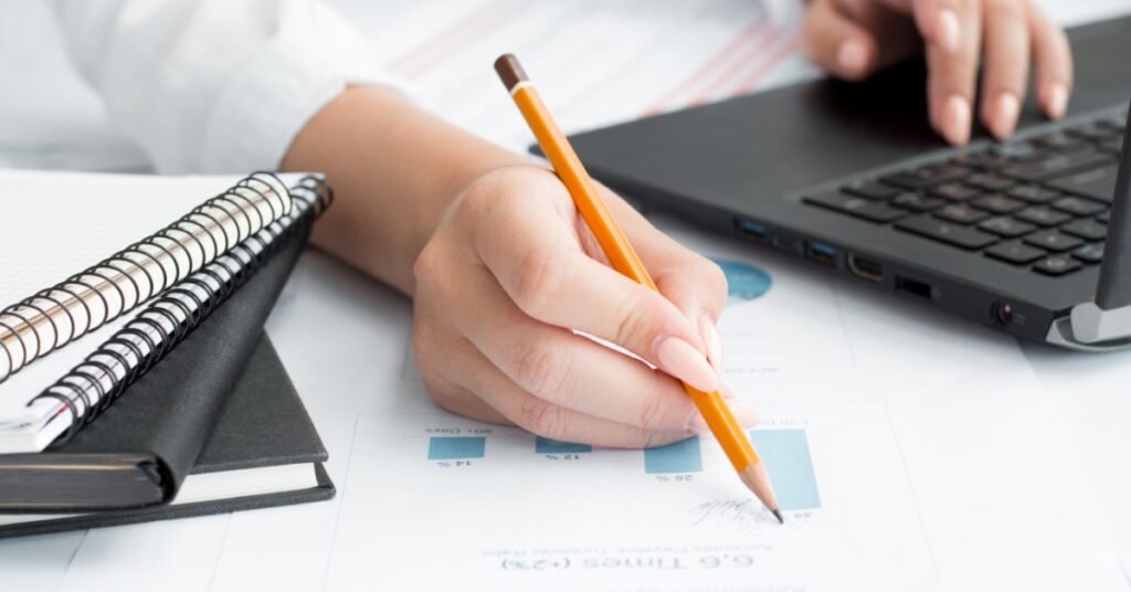 A close-up view shows a person holding a pencil up to a piece of paper with graphics on it. A laptop and notepads are next to it.