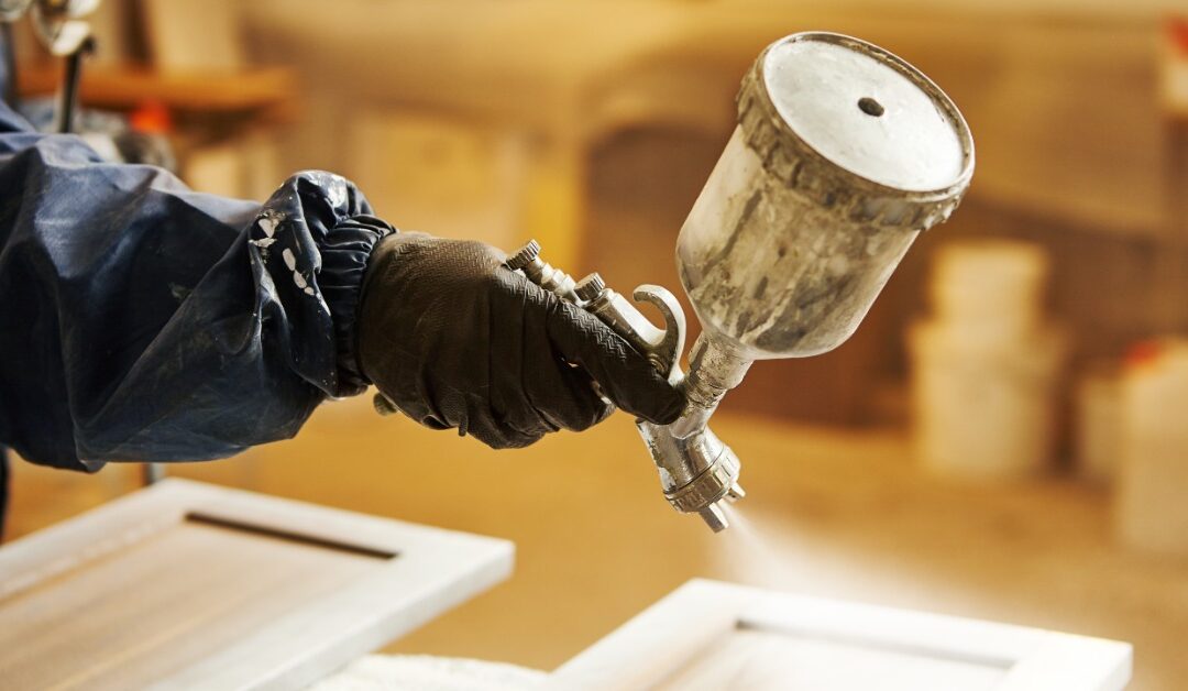 A person wearing a blue protective suit and protective face mask is using a sprayer to paint cabinet doors white.