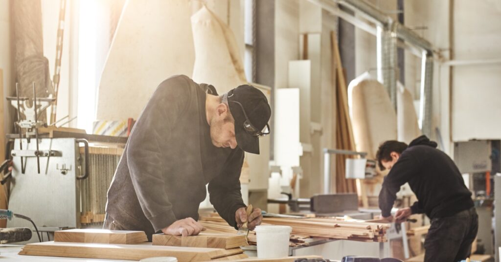 Two men wearing black hooded sweatshirts and dark jeans are working at workstations building cabinets.