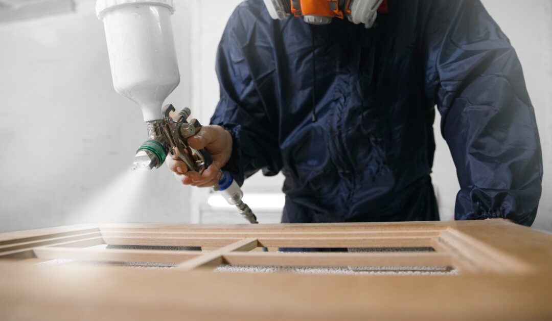 A close-up view shows a person wearing protective clothing and a gas mask while using a sprayer to paint cabinets.