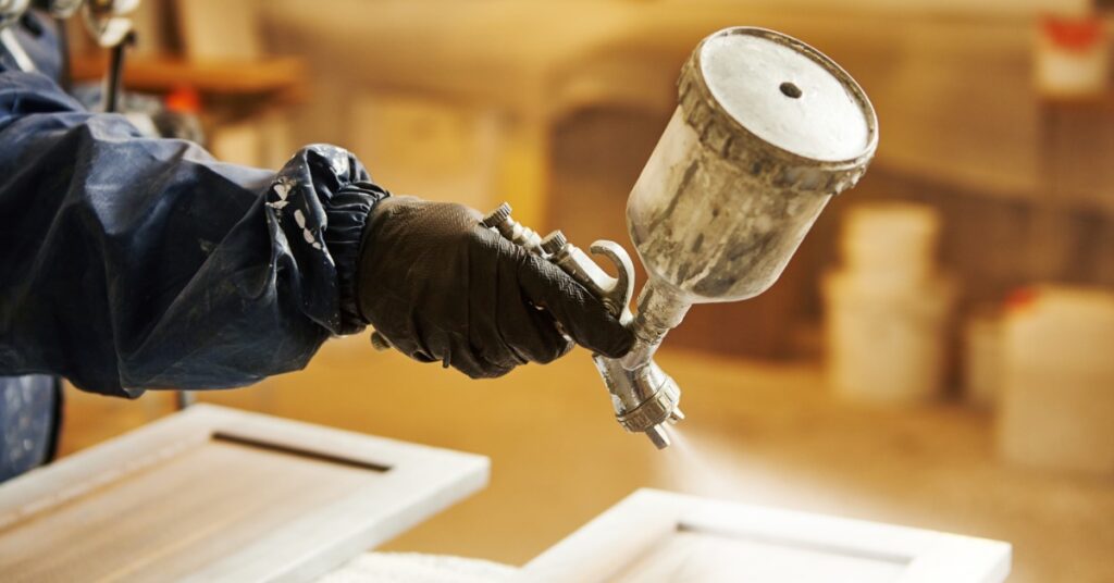 A person wearing a blue jumpsuit and a gas mask is using a sprayer to paint a cabinet door white.