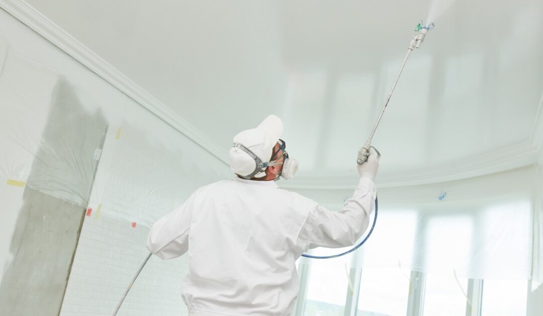 A man wearing white pants, a jacket, and a baseball cap is using a sprayer with a long nozzle to paint a ceiling white.