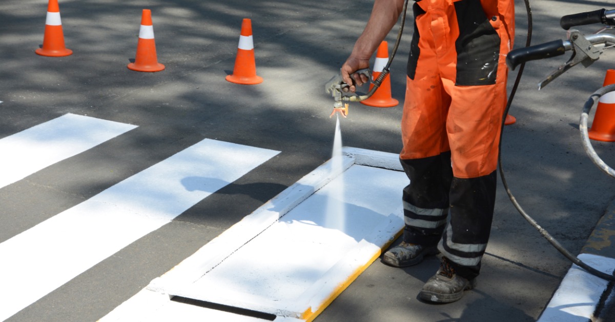 A man wearing bright orange pants is using a sprayer to paint a white rectangle on a road to create a sidewalk.