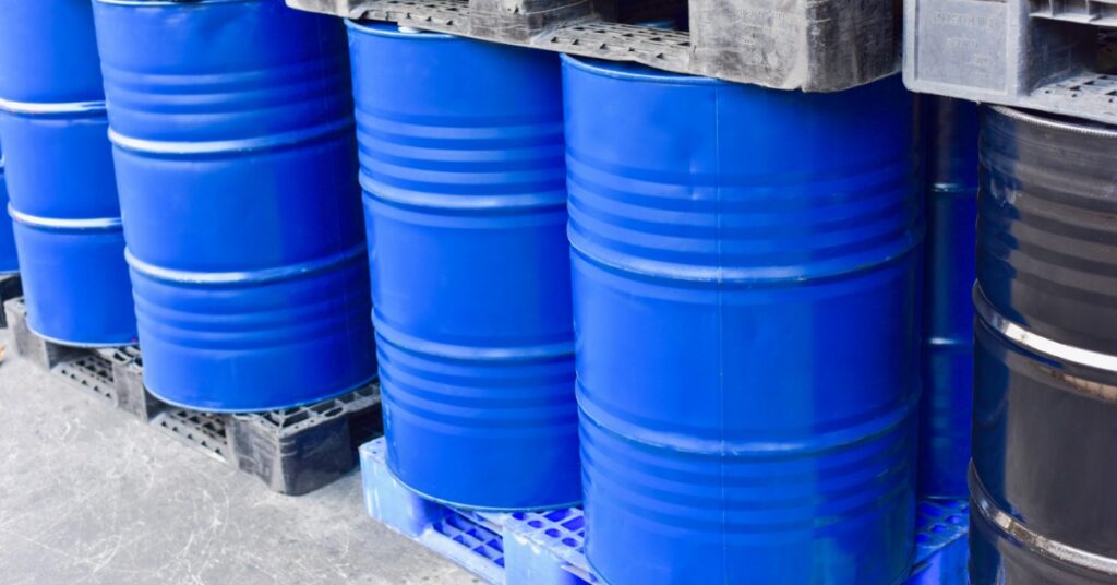 Large, blue metal drums sit on top of plastic pallets next to large, black metal drums. More drums are stacked on top.