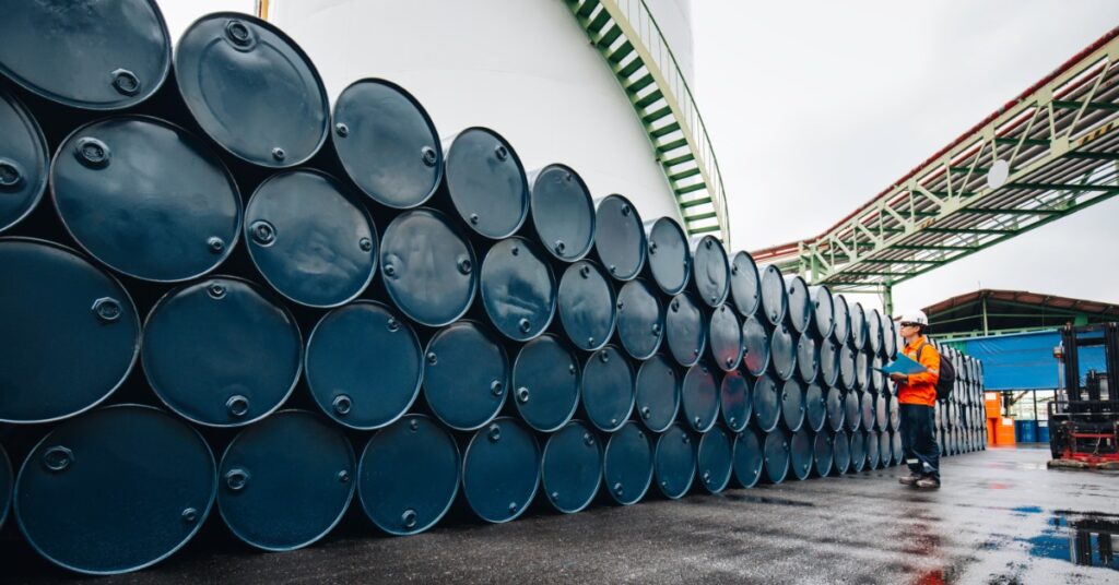 A large stack of blue barrels is stacked against an exterior wall. A man wearing a neon-orange jacket is looking at them.