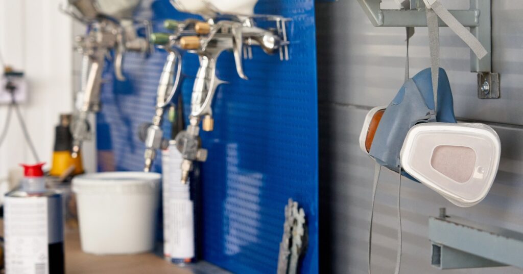 A close-up view shows a mask hanging on a metal hook. Industrial sprayers are hanging on hooks in the background.