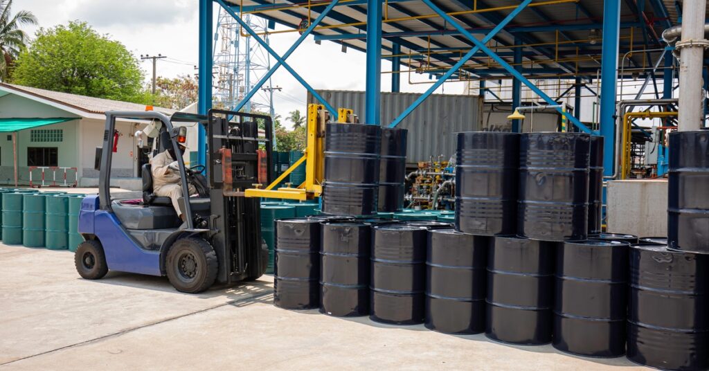 A person is using a blue forklift to move large black barrels outside. The barrels are stacked on top of one another.