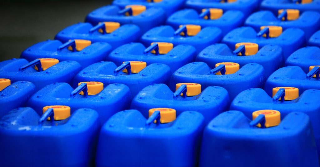 Four rows of blue plastic jugs with orange screw-top lids line up next to each other. The jugs have skinny handles.