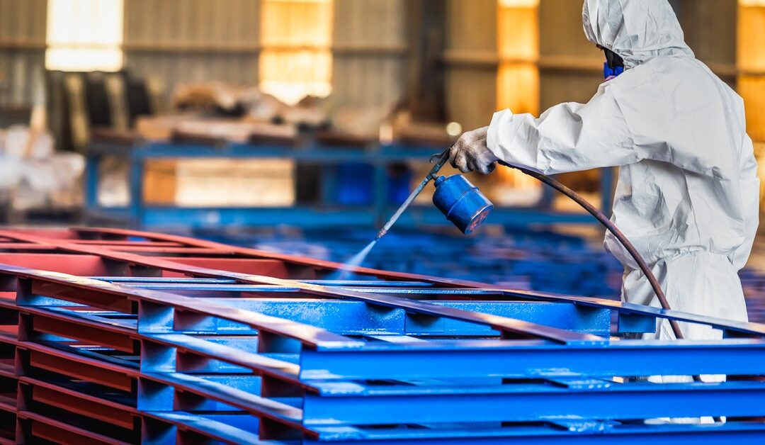A person wearing a white protective suit and mask is using an industrial sprayer to paint red steel beams blue.