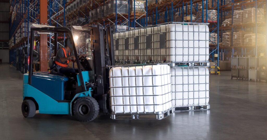 A person wearing dark-colored clothing is driving a forklift in a warehouse, moving a plastic storage container.