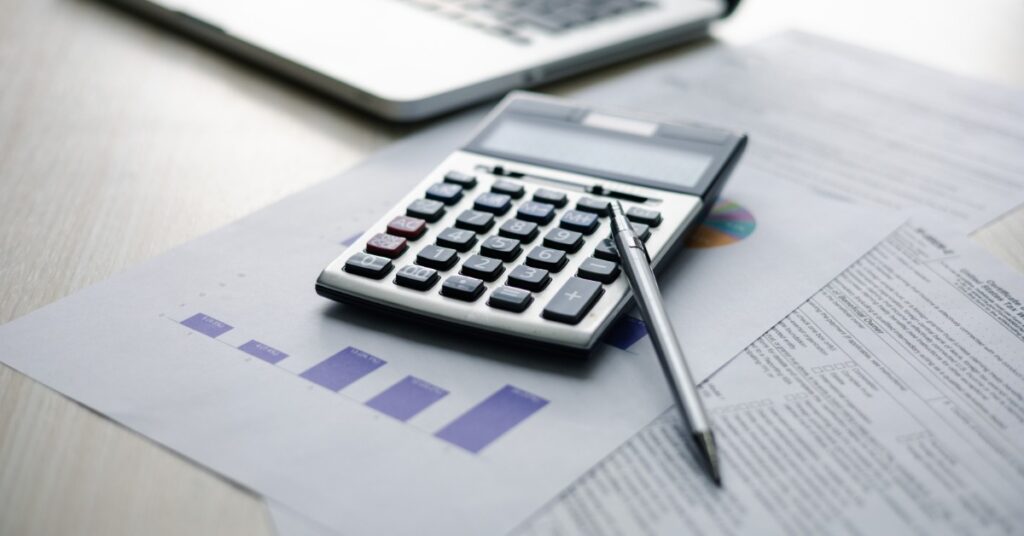 A small calculator sits on top of papers that are sitting on a desk next to a laptop. A pen sits on top of the calculator.