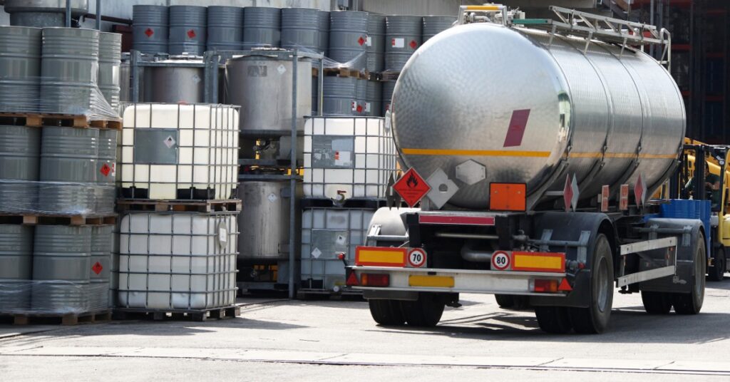 A tanker truck sits next to an organized collection of metal and plastic solvent containers that are outside.