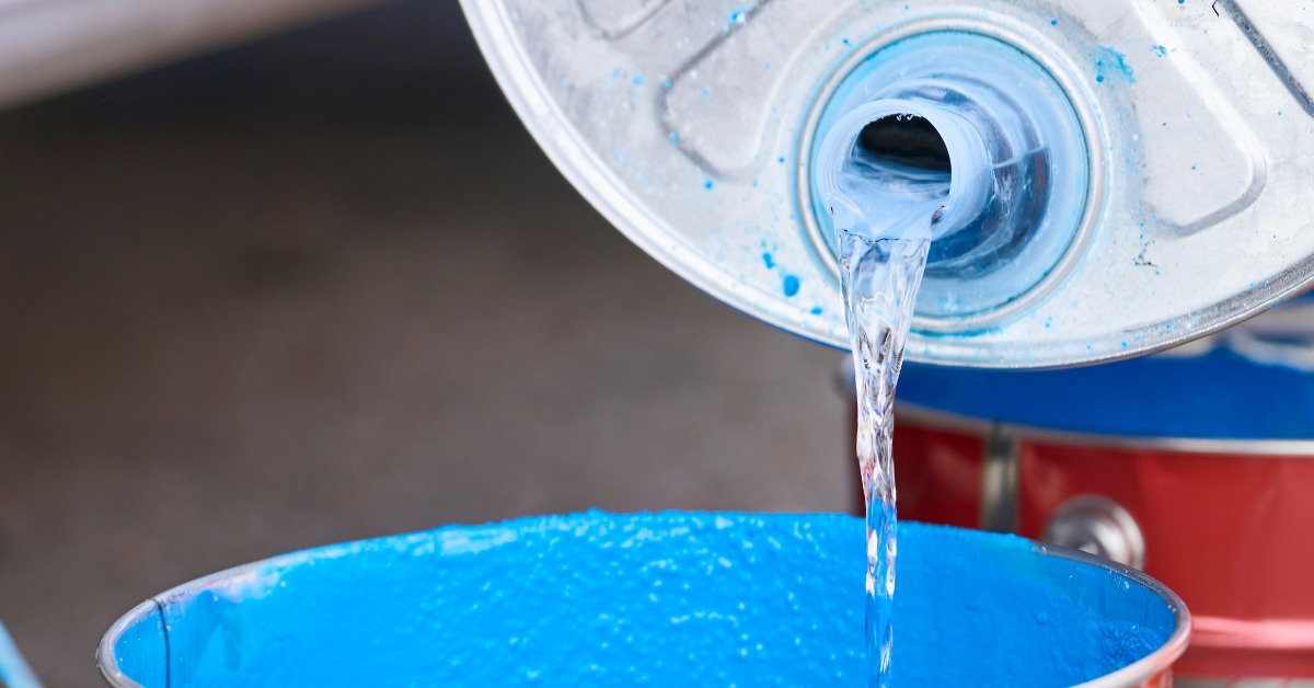 A person is pouring a clear liquid from a metal container into another metal container that's sitting on the ground.